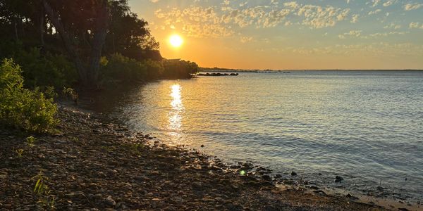 Sunset over a rocky shoreline with calm water and scattered clouds.