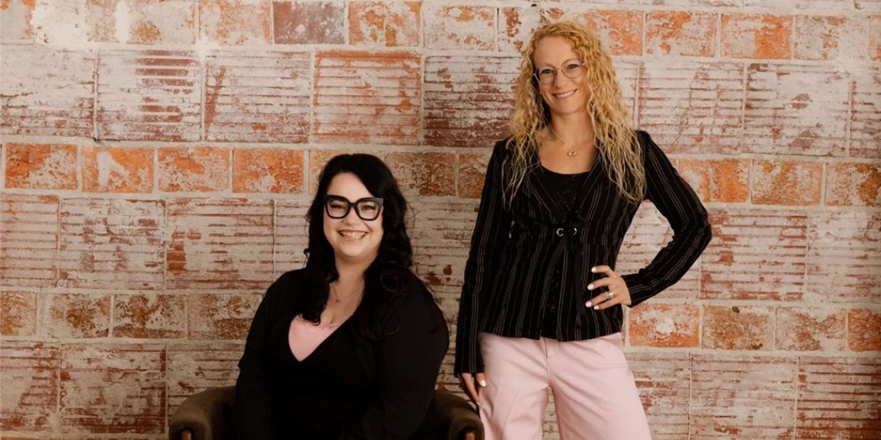 Two women smiling, one seated and one standing, in front of a textured brick wall.