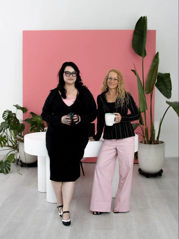 Two women standing by a white table with plants in a stylish indoor setting.