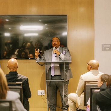 Man in suit speaking at a podium to an attentive audience in a conference room.