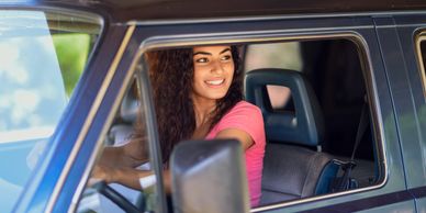 A smiling woman in a pink shirt sits behind the wheel of a car.