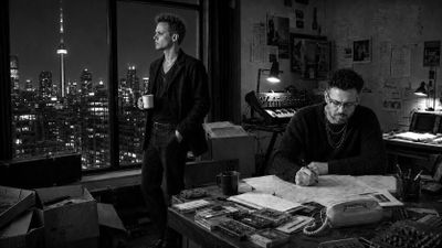 Two men in a dimly lit room with city skyline in the background.