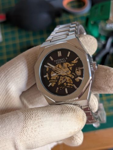 Close-up of a Thomas & George skeleton watch held by gloved hand.