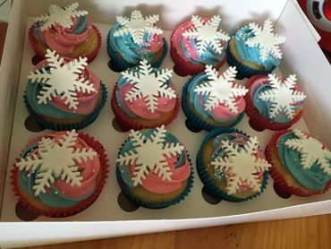 Box of cupcakes with pink and blue frosting topped with white snowflake decorations.