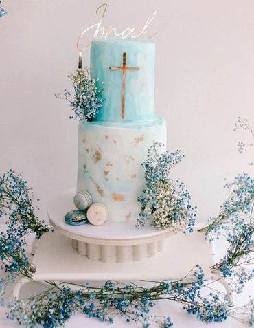 Elegant two-tier cake with blue and white hues, decorated with a gold cross and fresh flowers.