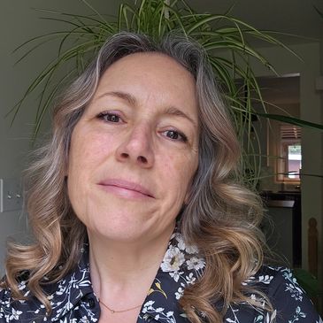 Woman with curled hair and floral shirt posing indoors with a plant behind her head.
