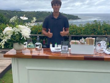Young man standing behind an outdoor bar with a scenic ocean view.