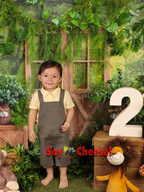 A toddler smiling in front of a leafy backdrop with a large number 2 and stuffed lion toy.