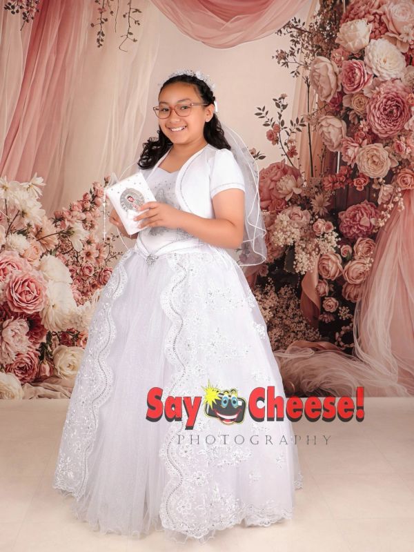 A young girl in a white dress holding a rosary and prayer book, smiling in front of floral backdrop.