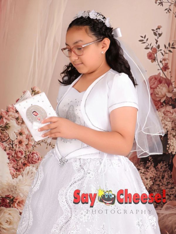 Young girl in a white dress reading a religious book with a floral background.