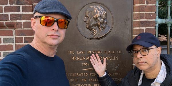 Two men pose beside a plaque marking Benjamin Franklin's last resting place.