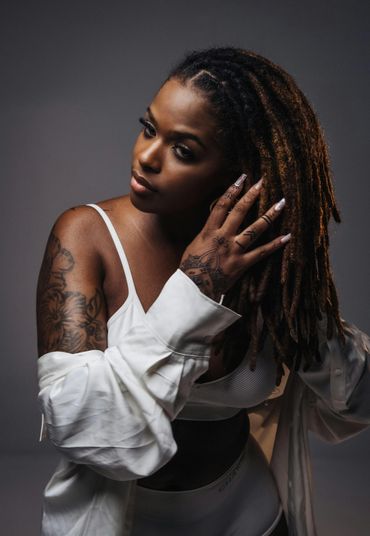 Woman with dreadlocks and tattoos adjusting her hair, wearing a white outfit.