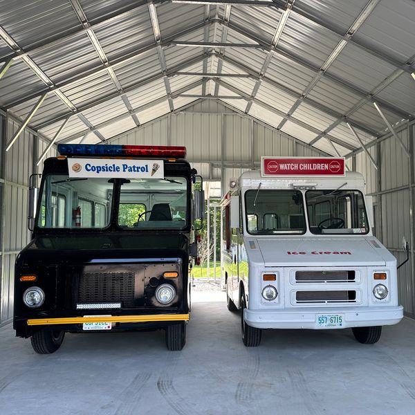 Two ice cream trucks parked inside a metal garage, one black and one white.