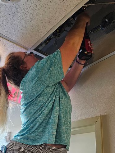 Woman on ladder fixing ceiling panel with power drill.