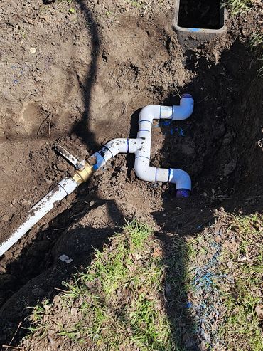 Outdoor plumbing with white PVC pipes and a valve in a dug-up patch of soil.
