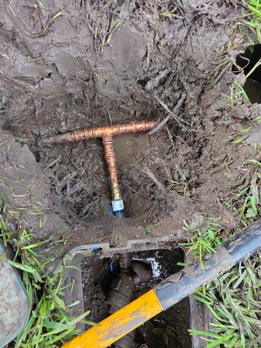 Open muddy ground with exposed copper water pipes and a tool.
