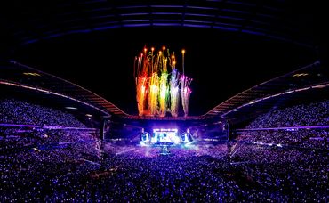 Nighttime stadium concert with vibrant fireworks and glowing crowd lights.