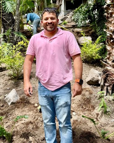 A smiling man in a pink shirt stands outdoors in a garden area.