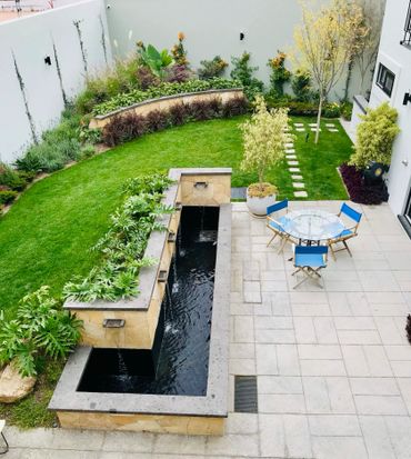 Modern backyard with water feature, green lawn, and blue chairs around glass table.