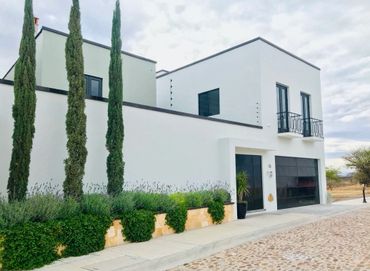Modern white house with tall trees and black doors under a cloudy sky.