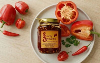 A jar of red pepper wine jelly surrounded by fresh red peppers on a plate.