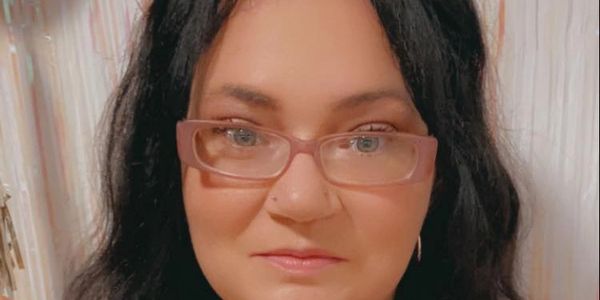 A woman with black hair and glasses smiles gently in front of a light, festive backdrop.