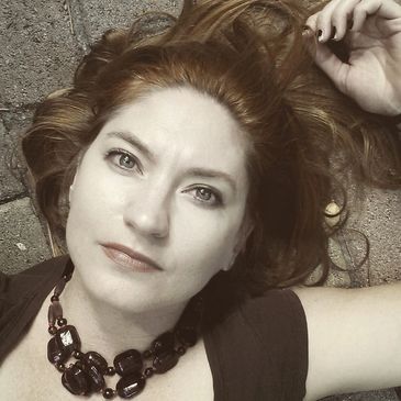 Woman with red hair lying down, wearing a dark necklace and top.