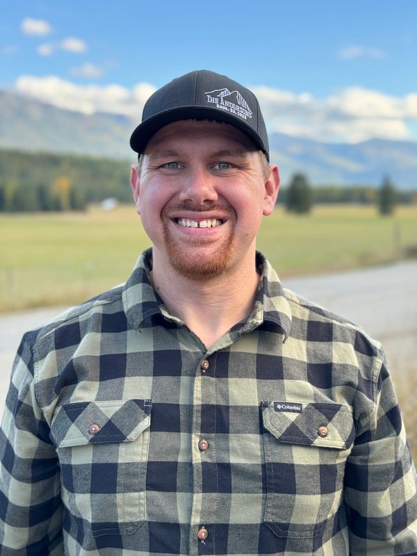 Smiling man in a checkered shirt and black cap standing outdoors with mountains in the background.