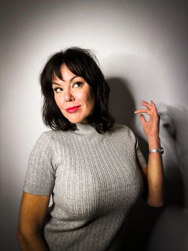 A woman with black hair and red lipstick poses against a plain wall.