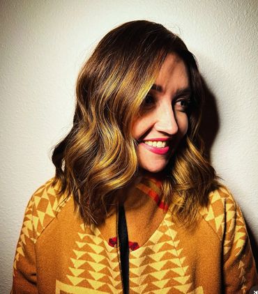 Smiling woman with wavy highlighted hair wearing a patterned brown coat.