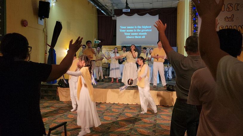 People worshiping with raised hands and singing on stage with lyrics projected.