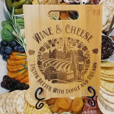 Engraved wooden cheese board with wine and cheese theme surrounded by crackers and fruits.