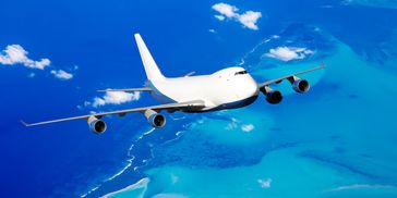 A white cargo plane flies over a turquoise ocean with visible coral reefs.