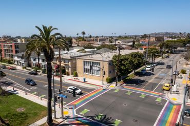 Rainbow crosswalk on a sunny urban intersection with palm trees.
