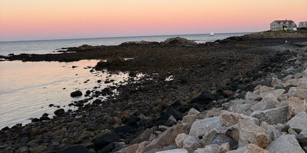 Rocky coastline at sunset with a pastel sky and a house in the distance.