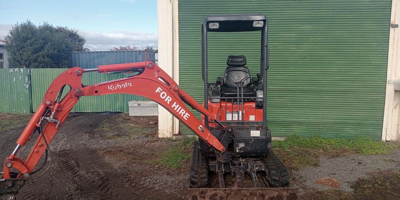 Small Kubota excavator with 'FOR HIRE' sign in front of green garage door.