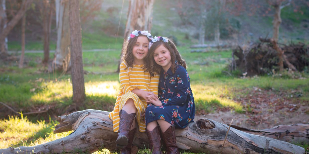 Sisters sitting on a log in the woods smiling for a photograph.