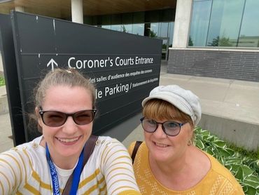 Two women smiling in front of a Coroner's Courts entrance sign.