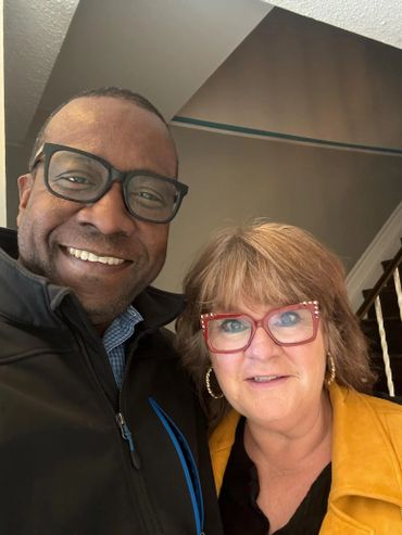 Two smiling people wearing glasses, posing indoors near stairs.