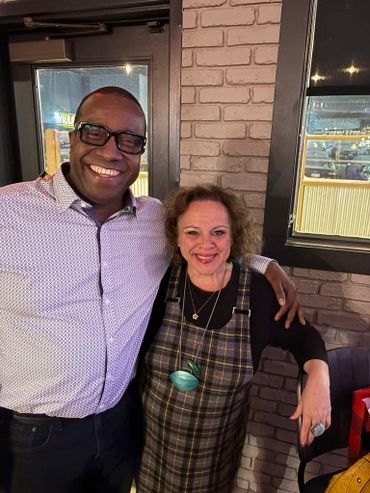 Two people smiling and posing together indoors with a brick wall background.