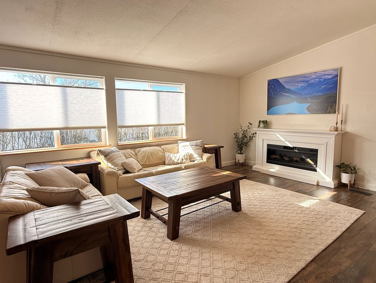 Bright living room with beige sofas, wooden tables, and a fireplace under a mountain landscape painting.