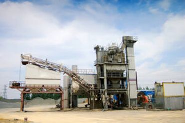 An industrial concrete or asphalt plant with large silos and conveyors under a blue sky.