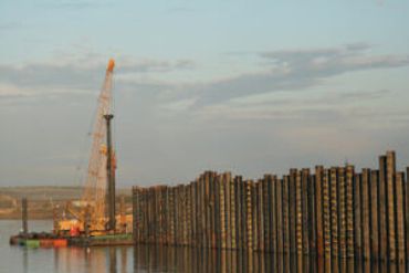 A pile driver works next to a tall wall of wood pilings at a construction site by a body of water