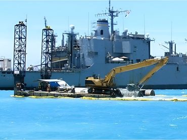 A yellow excavator on a floating platform dredges next to a large gray ship
