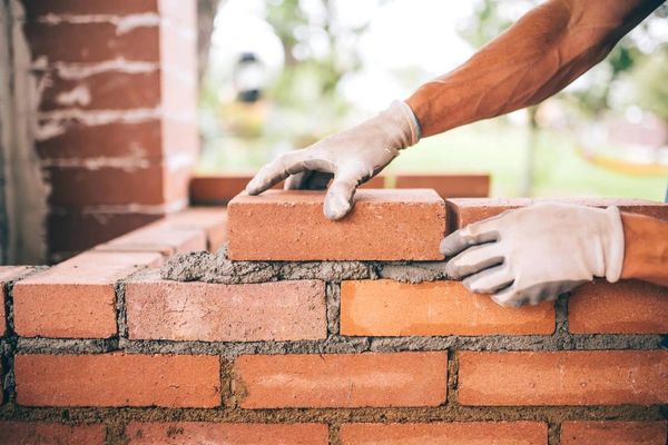 A close-up of a person's gloved hands laying a red brick on a mortar bed