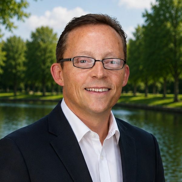 Smiling man in glasses and a suit outdoors by a water body.