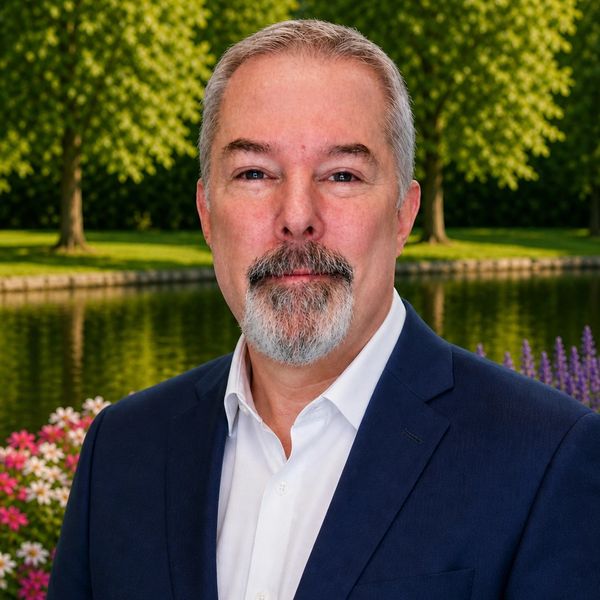 Mature man with gray beard in navy suit outdoors near a pond and flowers.