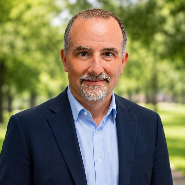 Middle-aged man in a blue blazer and light blue shirt, smiling outdoors.