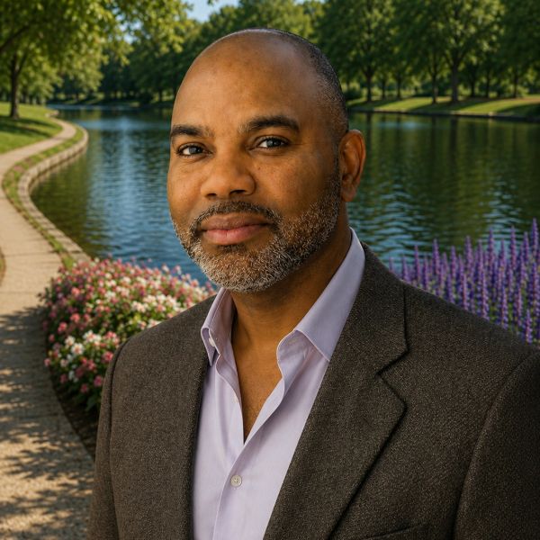 Confident man in a blazer stands by a scenic canal with flowers and trees.