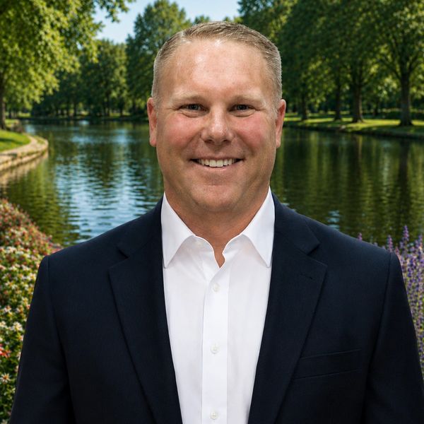 Smiling man in a suit stands by a serene lake with lush greenery.